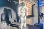 photo of a white spacesuit standing on stage at a conference, with a handful of onlookers in the background