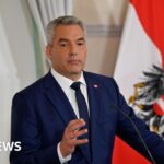 Karl Nehammer wearing a deep blue suit and a tie, standing in front of a microphone, with the Austrian and EU flags behind him