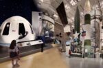 a white space capsule sits in a large museum hall, surrounded by other space artifacts